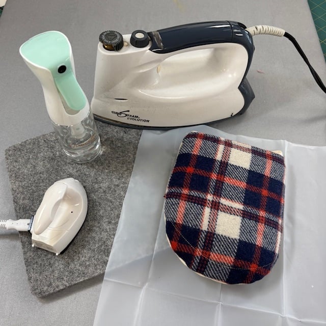 Essential pressing accessories for sewing including a mini iron, wool pressing mat, and mist bottle in a sewing workspace